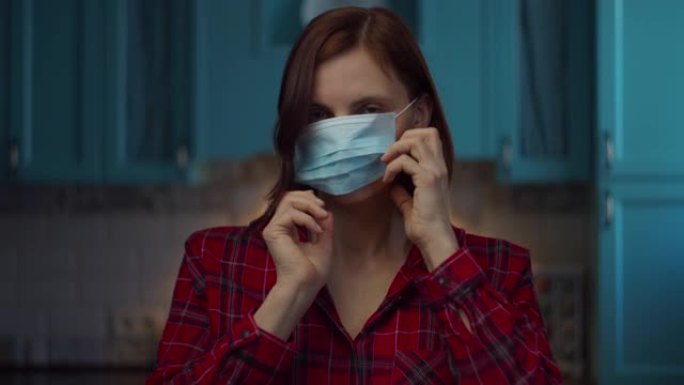 30多岁的年轻黑发女子脸上戴着医用口罩，微笑着看着相机。女性在医疗面具在家里的自我隔离从病毒。