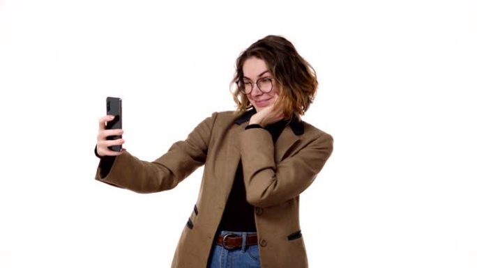 穿着棕色夹克的漂亮年轻女子在演播室里摆出自己的衣服，看着她的智能手机，在白色背景上自拍。做鬼脸