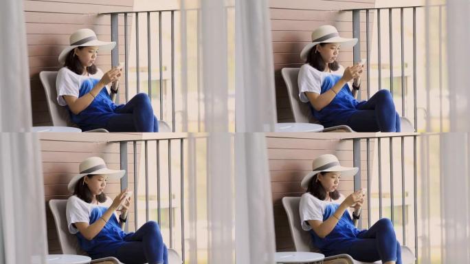 年轻漂亮的亚洲女人戴着宽帽子，使用智能手机坐在酒店房间阳台的椅子上。假日旅行概念。慢动作