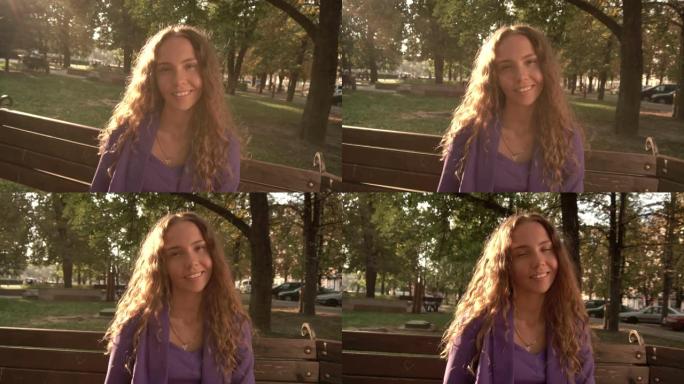 卷发的美丽女孩坐在公园的长椅上，看着相机。微笑的女孩，视频肖像，特写，慢动作