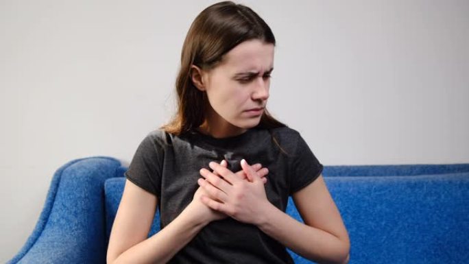 沮丧的年轻女子触摸胸部感到突然疼痛心脏病发作的胃灼热坐在家里的沙发上