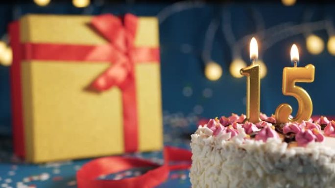 白色生日蛋糕编号15点灯燃烧的金色蜡烛，带灯的蓝色背景和用红丝带绑起来的礼物黄色盒子。特写