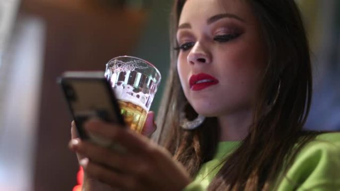 女孩晚上喝啤酒时看着手机屏幕。20多岁的迷人年轻女子在夜生活中检查智能手机
