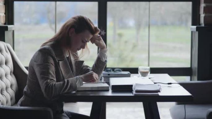 年轻的悲伤女孩由于工作的障碍和业务中的会计问题而坐着不高兴，坐在咖啡馆的桌子旁，用手握着头，一边在笔