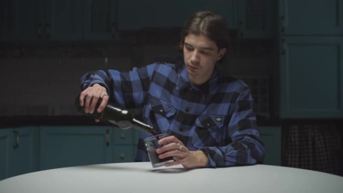 绝望的年轻人独自坐在厨房里喝红酒。心烦意乱的男性将红酒倒入玻璃杯中喝酒。