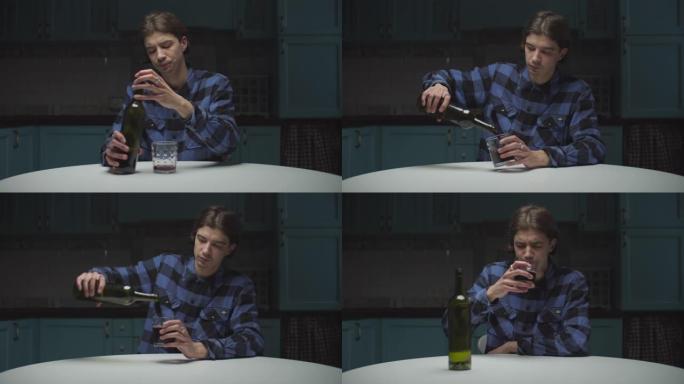 绝望的年轻人独自坐在厨房里喝红酒。心烦意乱的男性将红酒倒入玻璃杯中喝酒。