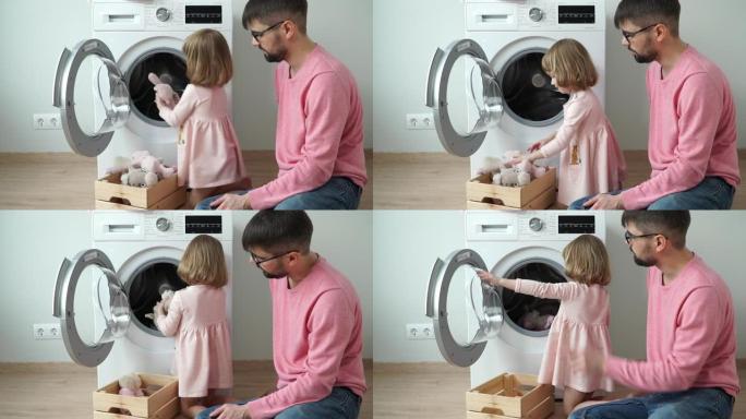 幸福的父亲肖像带着微笑的3岁女儿打开洗衣机的舱门，并放了柔软的玩具进行洗涤。家用概念和家用电器