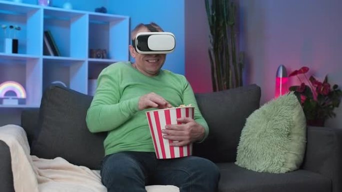 高个子白人男子使用虚拟现实耳机观看视频，坐在沙发上吃爆米花在房间里