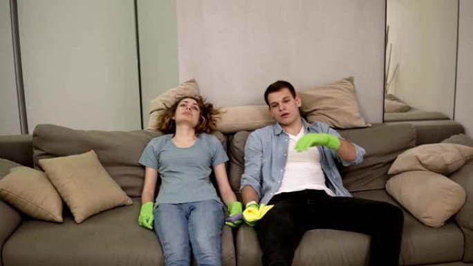 筋疲力尽的白人年轻夫妇在清洁后倒在沙发上，清洁后感到疲倦，额头上挥出汗水。呼气。戴着绿色橡胶手套。慢