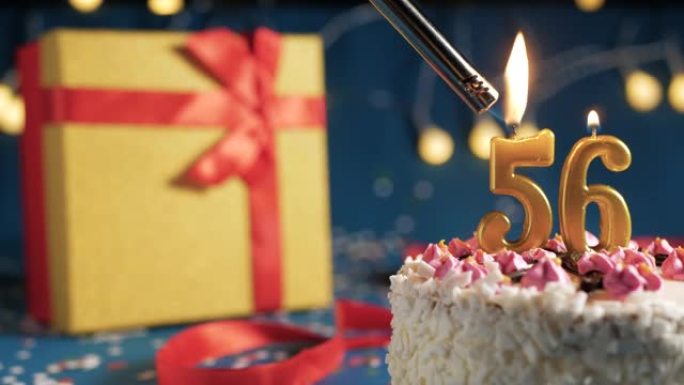 白色生日蛋糕56号点火器燃烧的金色蜡烛，蓝色背景带灯和用红丝带捆绑的礼物黄色盒子。特写
