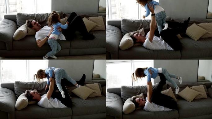 幸福的家庭父亲和小女儿一起躺在沙发上玩得开心父亲发痒女孩，举起他的怀抱，她笑了。慢动作