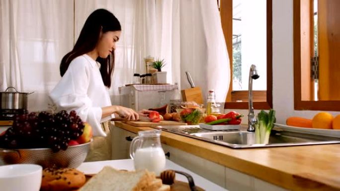 年轻的亚洲可爱女人在厨房切碎西红柿，早上桌子上有水果，牛奶罐和面包。
