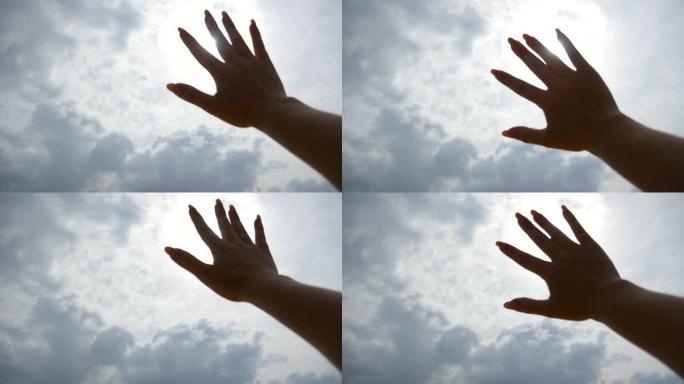 明亮的阳光穿透了年轻女子伸出手臂的手指，在多云的天空背景下。女性手的特写伸向太阳。暑假或假期的概念。
