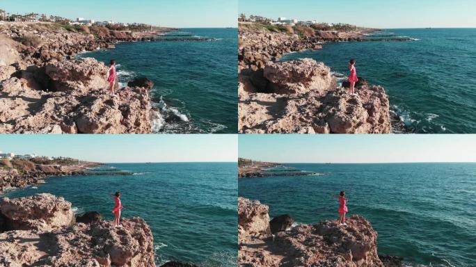 山崖顶上穿着红色连衣裙的女人，双手分开。当她享受阳光和暖风时，风拍打着她的衣服。手臂分开和海浪碰撞的