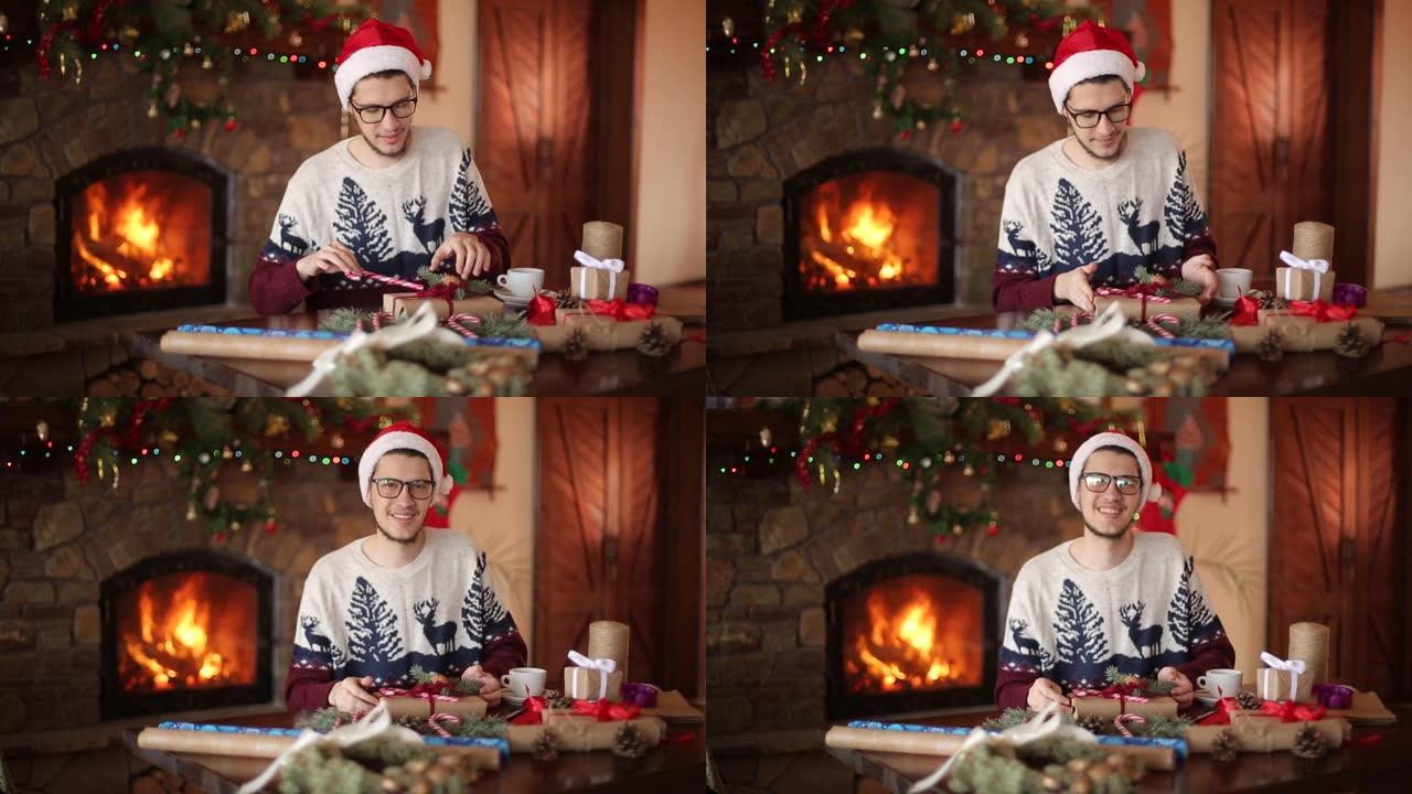 大胡子的男人坐在壁炉附近的新年礼物上鞠躬，看着镜头。戴着圣诞帽的家伙用纸包装礼物盒子，增加了冷杉树枝