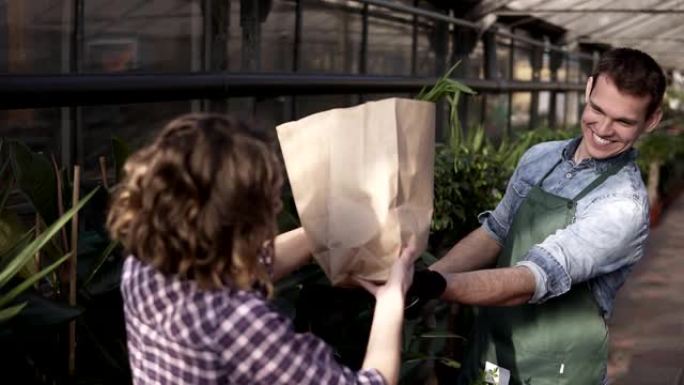 穿着绿色围裙的积极高大的推销员在温室市场向顾客提供纸袋中的新鲜蔬菜。微笑的男人向女顾客推销蔬菜。人与