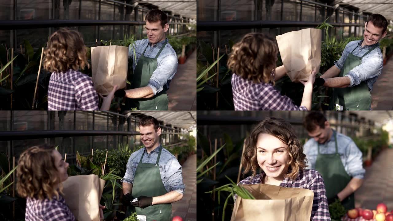 穿着绿色围裙的积极高大的推销员在温室市场向顾客提供纸袋中的新鲜蔬菜。微笑的男人向女顾客推销蔬菜。人与