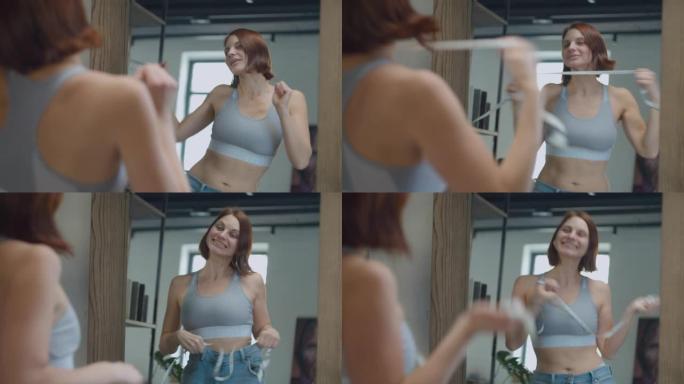 30多岁的年轻女性穿着大尺寸的旧牛仔裤欣赏减肥的结果。快乐的女性用厘米胶带在镜子里跳舞