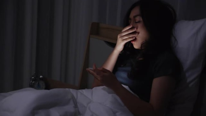 亚洲年轻女子躺在床上，晚上在卧室里关掉灯玩智能手机。在手机上阅读社交。在弱光下使用手机对眼睛有健康影