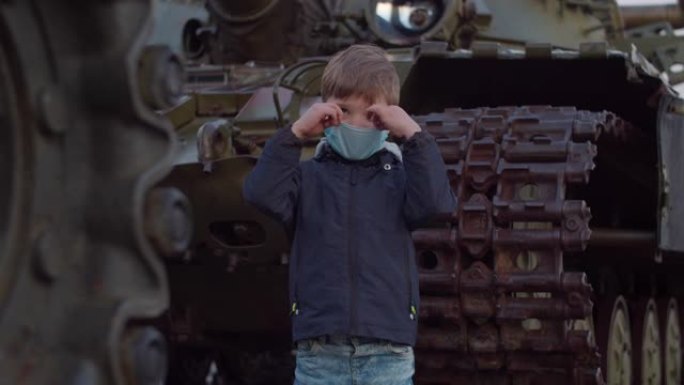 在冠状病毒新型冠状病毒肺炎大流行期间，戴着医用防护面具的男孩站在军用机器旁，四处观察。检疫国家紧急状