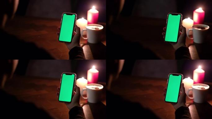使用绿屏智能手机的人的肩视图。在木桌上模糊蜡烛灯和一杯咖啡。