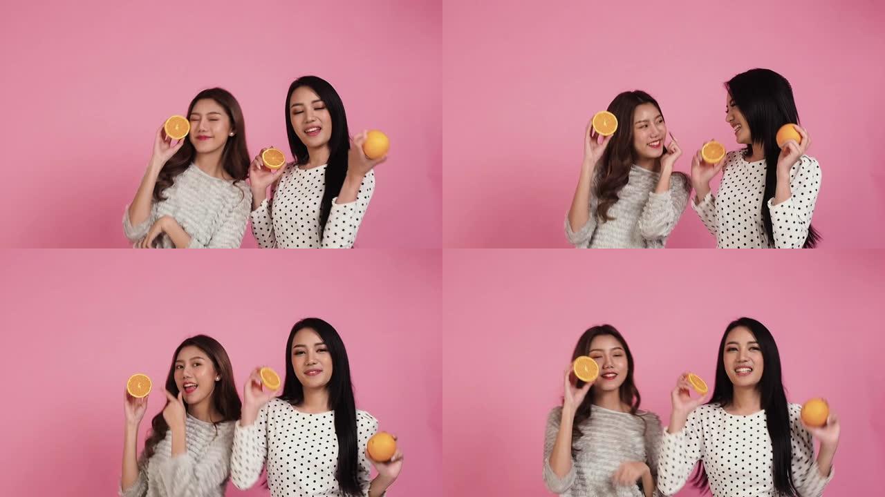 幸福两个迷人的女人在粉红色的背景下跳舞。亚洲美女拿着切片橙色水果健康cear概念