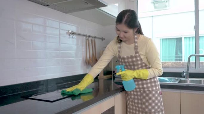 亚洲妇女戴着橡胶保护的粉红色手套，拿着抹布和喷雾瓶洗涤剂，双手放在臀部，观察干净和闪闪发光的厨房。家