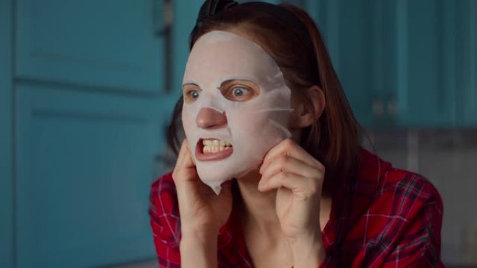 有趣的30多岁女人脸上戴着白色化妆品面膜在家做鬼脸。女人在蓝色厨房的面具上做鬼脸。