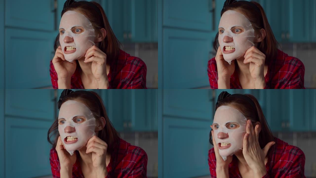 有趣的30多岁女人脸上戴着白色化妆品面膜在家做鬼脸。女人在蓝色厨房的面具上做鬼脸。