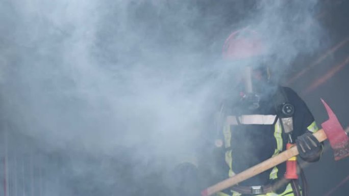 亚洲消防员手持斧头自信地穿越烟雾，背景中燃烧着烈火。消防员生涯，训练消防演练理念。慢动作。