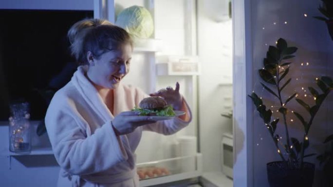 夜间饥饿，年轻的女孩在厨房里穿着睡袍打开冰箱，急切地咬了一个汉堡，然后手里拿着盘子离开房间