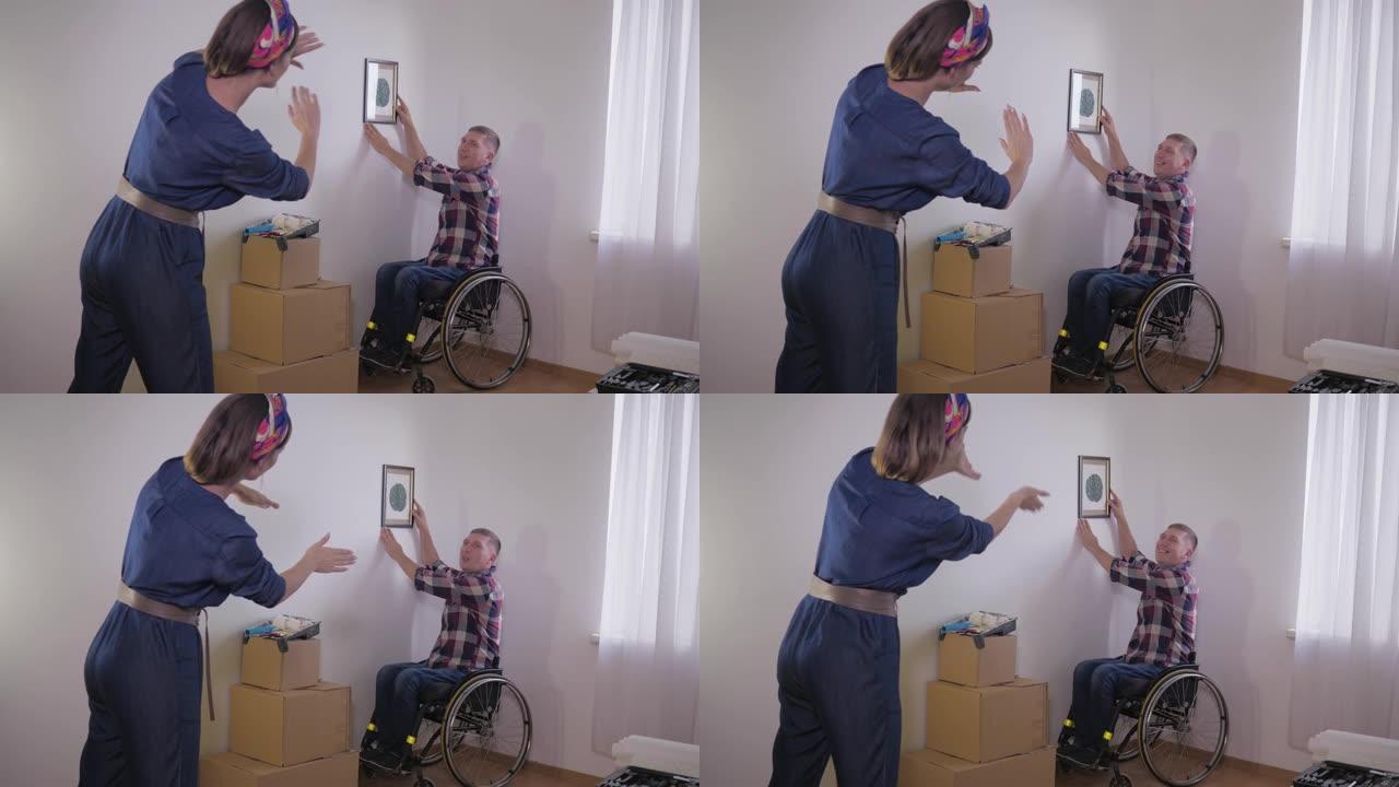 坐在轮椅上的乐于助人的残疾人悬挂照片，而他的妻子用手指做框架，并选择在白色墙壁上拍照的地方