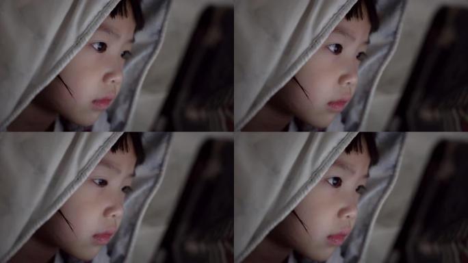 可爱的亚洲小女孩躺在毯子下晚上在卧室看数字笔记本电脑。手持镜头，真实直播