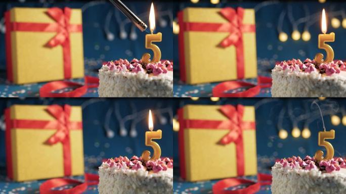 白色生日蛋糕5号点火器燃烧的金色蜡烛，蓝色背景带灯和用红丝带捆绑的礼物黄色盒子。特写
