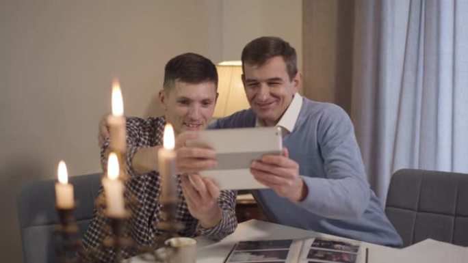 高加索成年父子拥抱并在平板电脑上自拍的肖像。快乐的中年男人和开朗的家伙晚上一起在家休息。生活方式、休