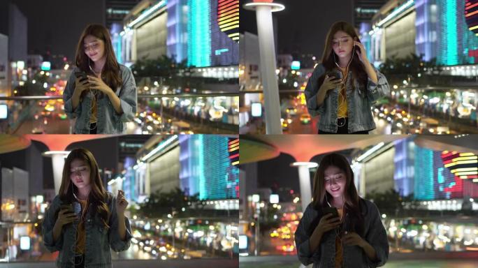 4k年轻美丽的亚洲女人带着耳机散步，晚上在城市里听智能手机播放在线音乐。十几岁的女孩在城市之夜享受广