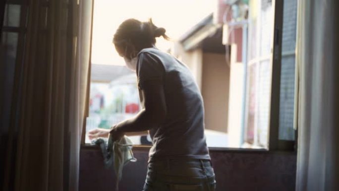 年轻的亚洲女性在晚上用日落背光背景和面罩保护清洁窗户。做家务概念。
