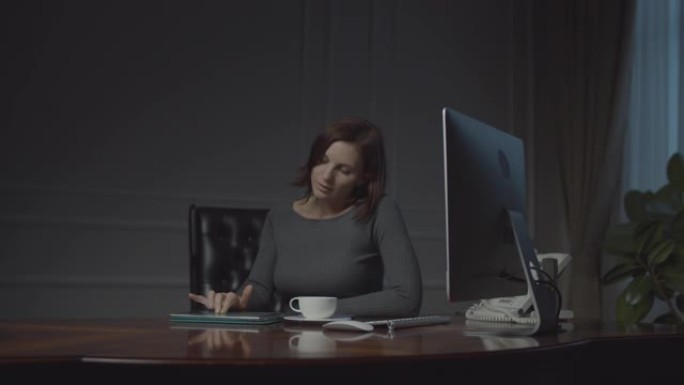 年轻的女商人晚上在商务办公室工作平板电脑。坐在舒适的办公室皮革扶手椅上喝咖啡的女人。
