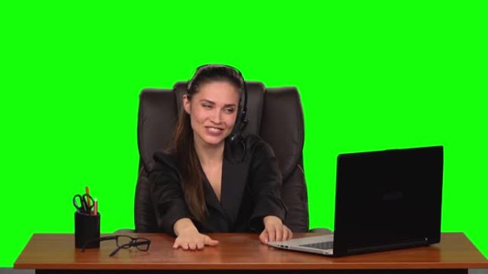 戴着耳机的商务女孩用网络摄像头说话，带着喜悦的心情看着笔记本电脑。绿屏。慢动作