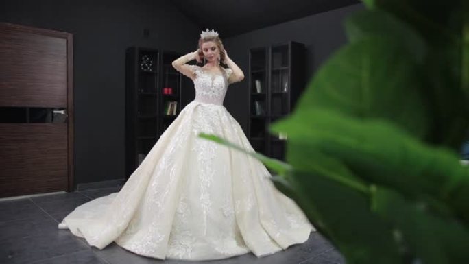 年轻的金发新娘穿着白色婚纱，戴着皇冠，在房间里为相机摆姿势