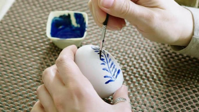 画复活节彩蛋的女人特写。女性的手在鸡蛋上画了蓝色的花卉图案，彩绘成白色。4K