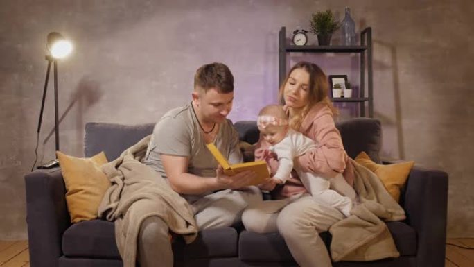 一个年轻的家庭坐在舒适房间的沙发上。妈妈、爸爸和宝宝看书。家庭快乐