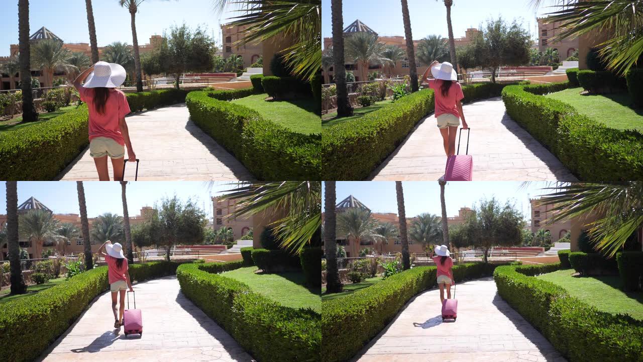 穿着白色大帽子和粉色旅行包的美女走在酒店的地盘上。她走在有棕榈树的小巷上。1920x1080中的高清