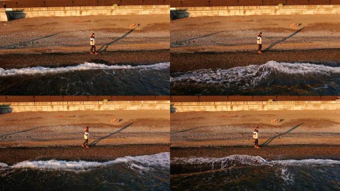 鸟瞰图年轻女孩摄影师带着相机在日落的黄金时刻走在海滩上。美丽的海浪在夕阳的光芒中袭击了海岸。独立海上
