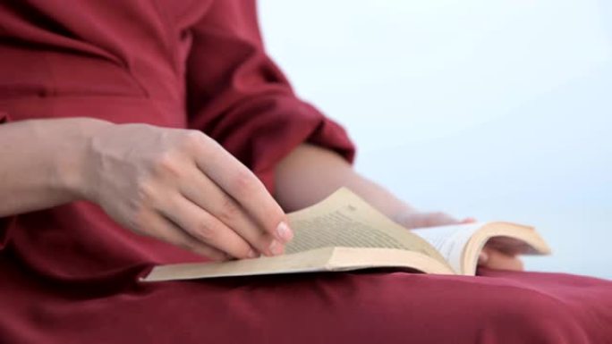 特写镜头一位穿着红色鲜艳连衣裙和草帽的迷人高加索女孩在日落时坐在海边的一块大石头上，读一本有趣的书