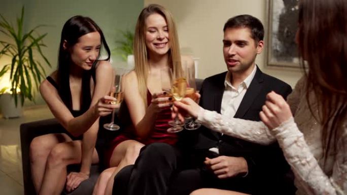 追踪快乐自信的年轻人坐在沙发上，与三个漂亮的女人调情，在家庭聚会上喝酒的中等镜头