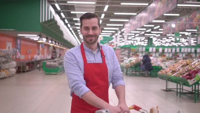 超市里快乐微笑英俊的中年卖家男人的肖像带着购物车积极的千禧一代员工看着相机，笑脸满足于好工作成功的人