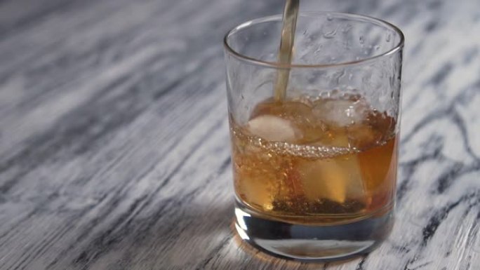将金色威士忌倒入一个装有冰块的玻璃杯中，放在一个旧的木制灰色酒吧柜台上