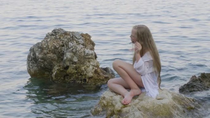 穿着白色外衣的少女坐在夏日海滩海水中的岩石上。穿着比基尼的女摄影师拍摄女孩少年在水中的海石上。