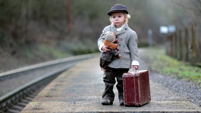 火车站上的可爱男孩，带着手提箱和漂亮的老式娃娃等着火车